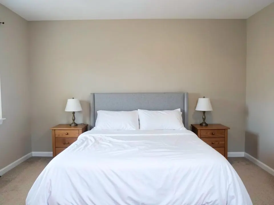 AI-generated example: plain beige bedroom seen straight on, a gray upholstered headboard against a bare beige wall, white bedding, two wooden nightstands with lamps, white baseboards and beige carpet