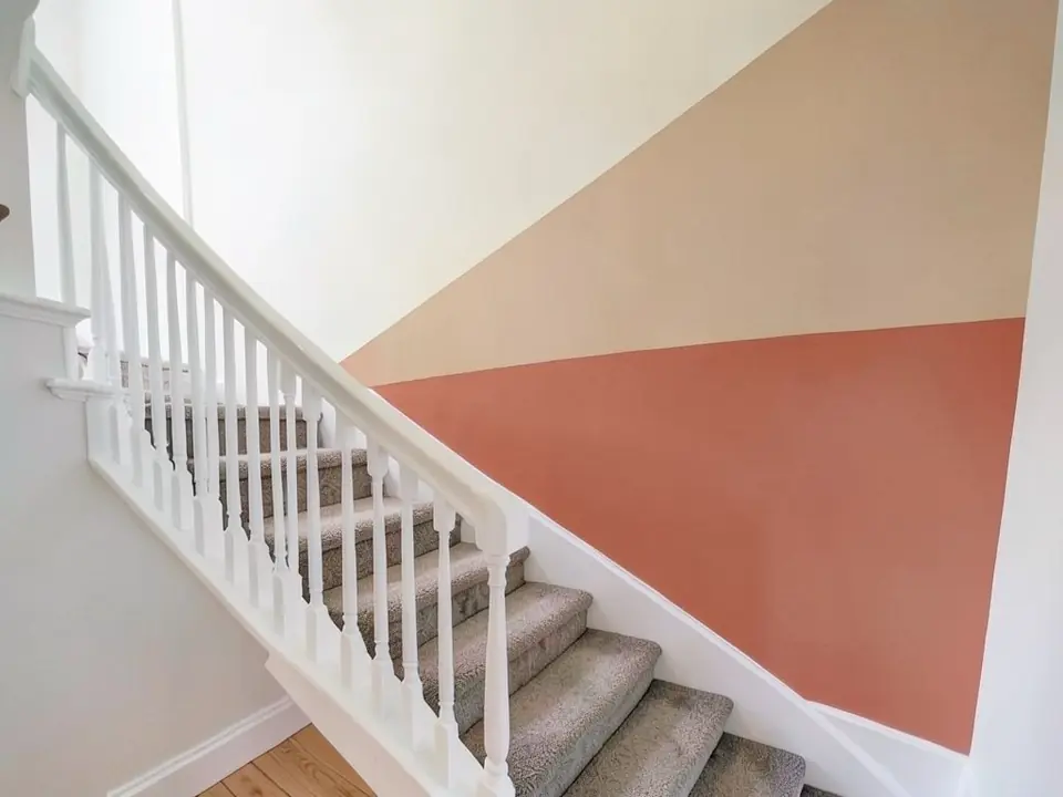 AI-generated edit of the same staircase: the tall wall now has terracotta on the lower part and sand beige above it, split by a nearly level line, with a white triangle left in the upper-left part of the wall; stairs, carpet, railing and the left wall unchanged