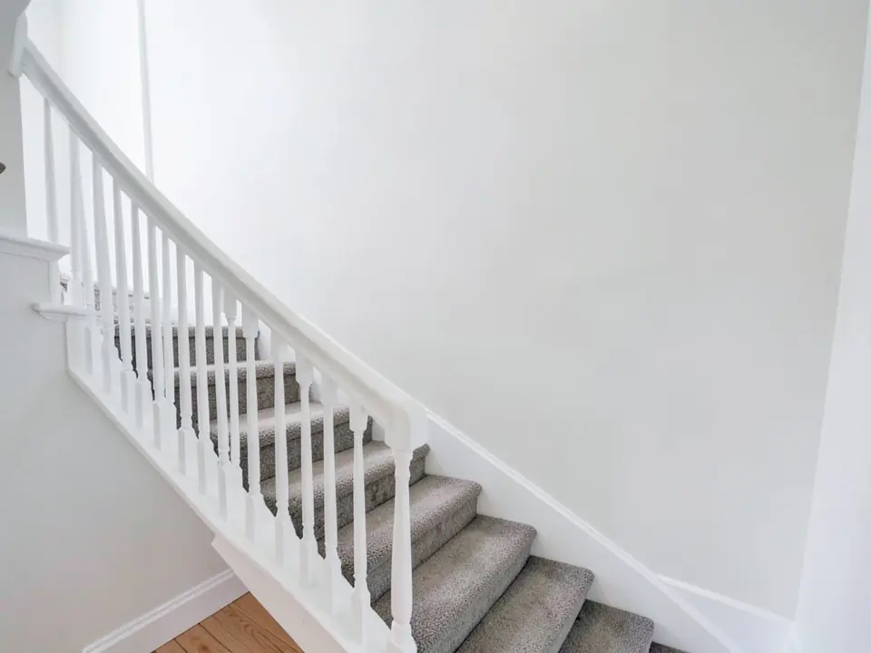AI-generated example: carpeted staircase with a white handrail and white balusters running up beside a tall, bare white wall