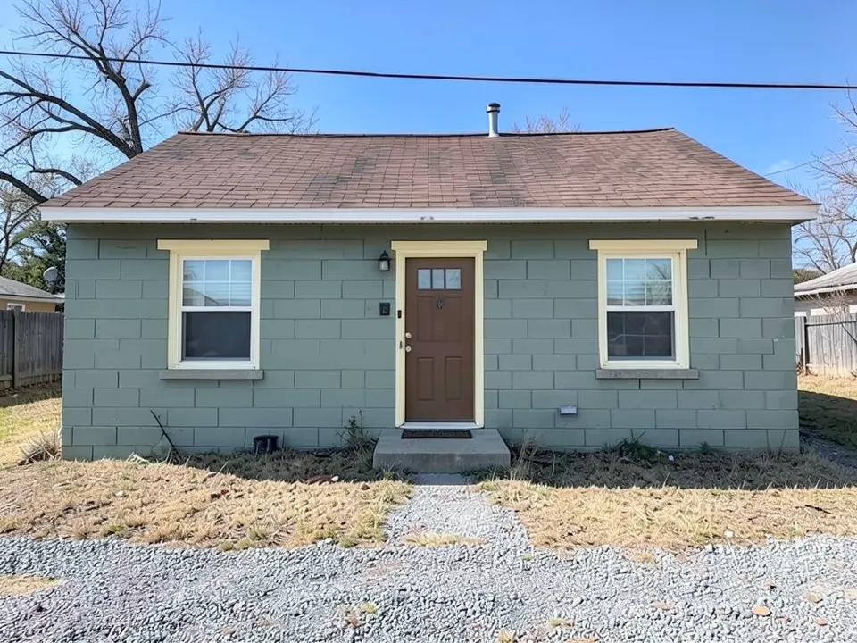 AI-generated edit of the same house: every block wall is now an even muted gray-green with the mortar lines showing, and the window and door trim is a pale yellowish cream; the brown door, windows, gray sills, roof, flue pipe, step, gravel, grass and trees are kept