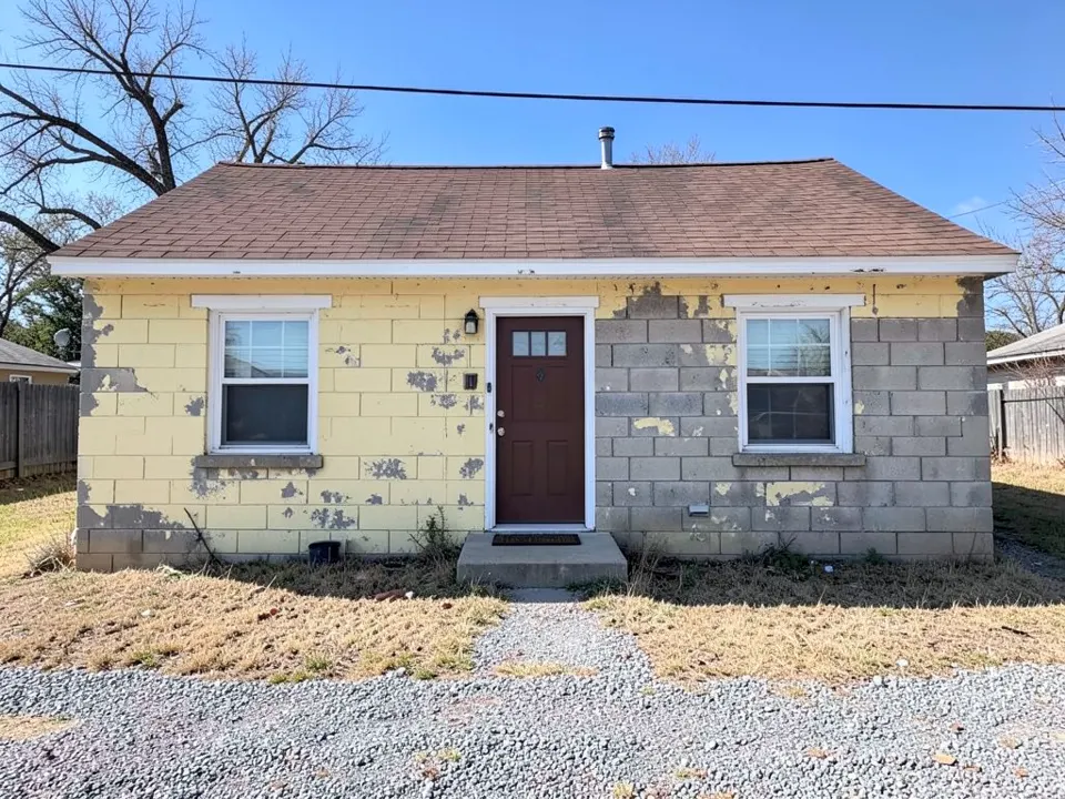 AI-generated stand-in: small single-story concrete block house with faded pale yellow paint peeling off the walls in patches of bare gray block, white window and door trim, a brown front door, a brown shingle roof with a metal flue pipe, a concrete step, dry grass, gravel and bare trees