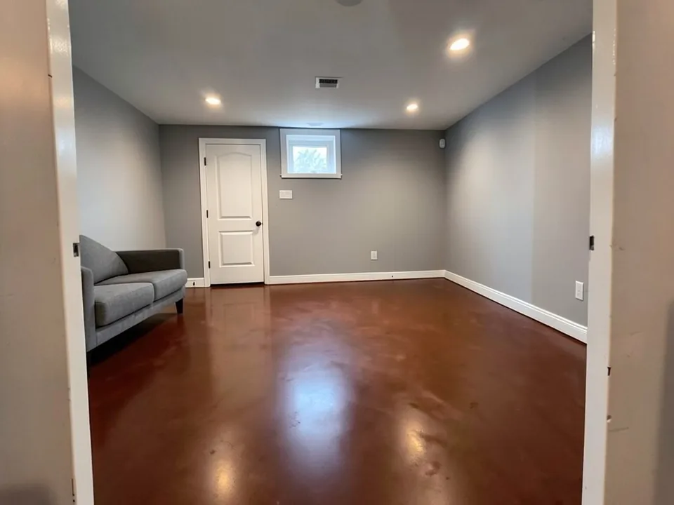 AI-generated edit of the same basement: the floor is now a reddish coffee-brown with soft mottling and glossy reflections of the ceiling lights, and the scuff marks and arc lines are gone; the walls, baseboards, door, window, sofa and ceiling are kept, with the view shifted slightly