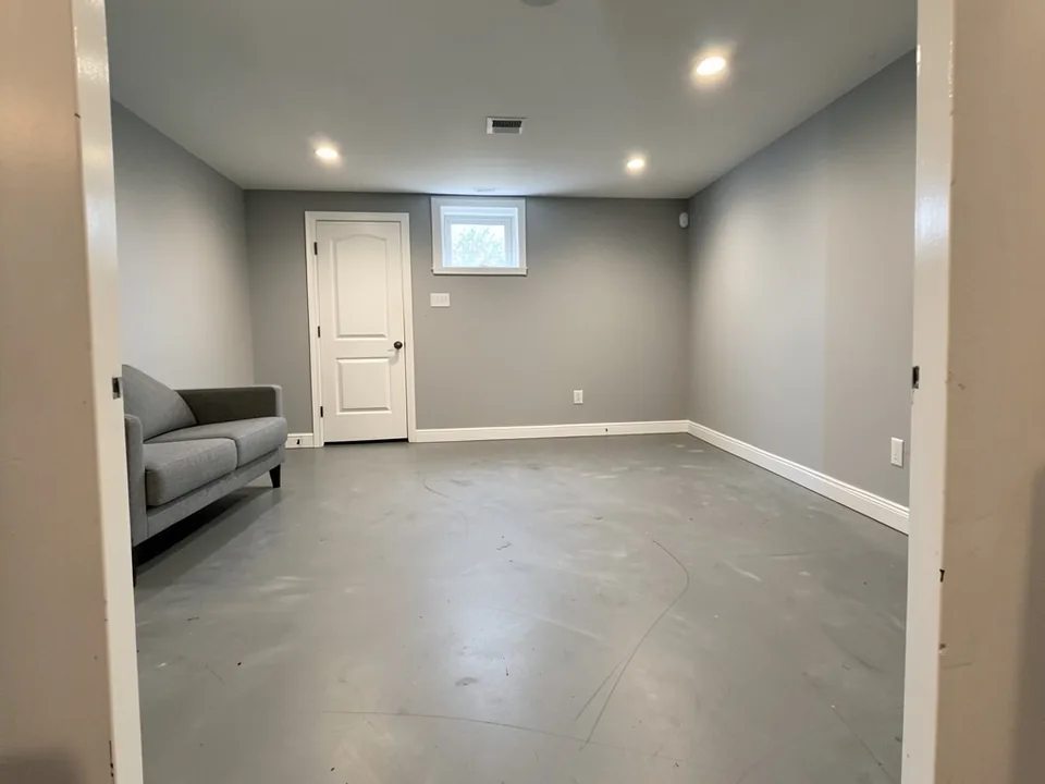 AI-generated example: basement room with a plain gray concrete floor showing scuff marks and faint arc lines, gray walls, white baseboards, a white door, a small window, recessed ceiling lights and a gray sofa on the left