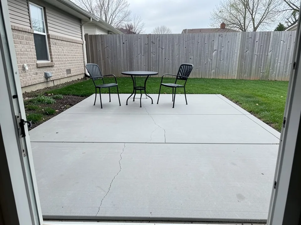 AI-generated example: plain gray broom-finished concrete patio seen through an open back door, with saw-cut joints, two hairline cracks, two black metal chairs and a round table, a lawn, a wooden privacy fence and a brick and siding house wall