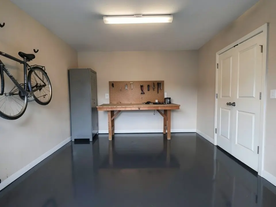 AI-generated edit of the same garage: the floor is now one even solid charcoal epoxy in high gloss, reflecting the workbench; bicycle, cabinet, doors and white baseboards unchanged