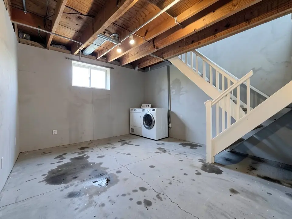 AI-generated example: unfinished basement with a gray concrete slab covered in dark water stains and hairline cracks, a white staircase, a washer and dryer and exposed ceiling joists