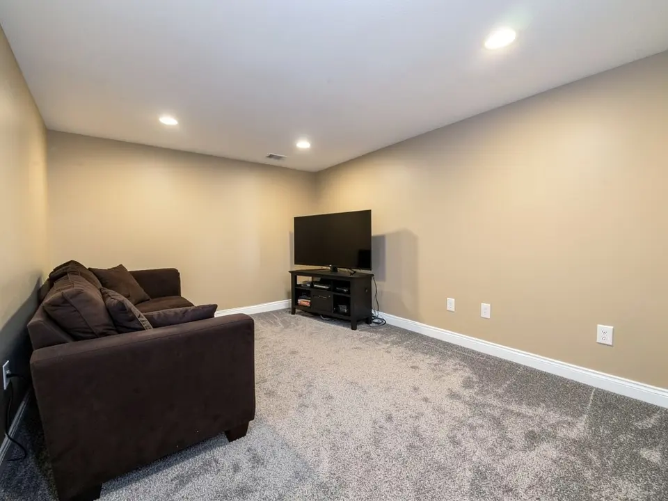 Finished basement room with beige walls, white baseboards, gray carpet, a dark brown sofa, a TV on a stand and recessed ceiling lights, before