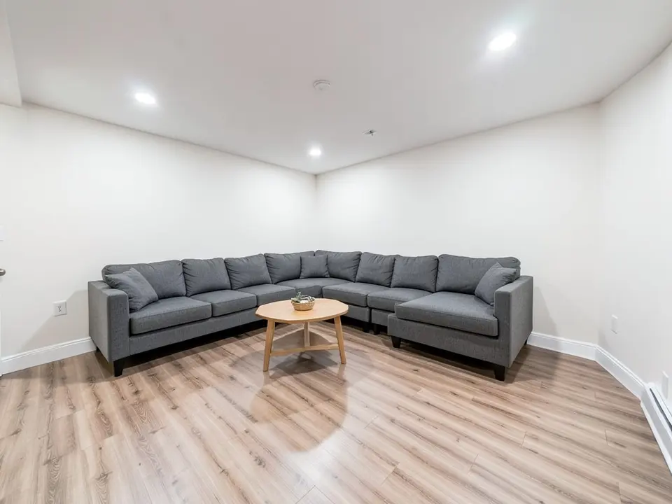 Finished basement room with white walls, white baseboards, a light wood-look floor, a gray sectional sofa, a round wood coffee table and recessed ceiling lights, before