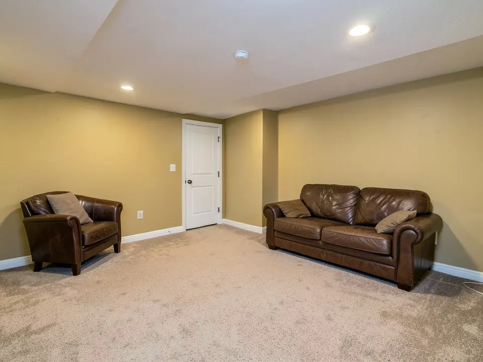 Basement family room with dull yellowish-beige walls, white baseboards, beige carpet, a brown leather sofa and armchair, a white paneled door and a white ceiling with a recessed light, before