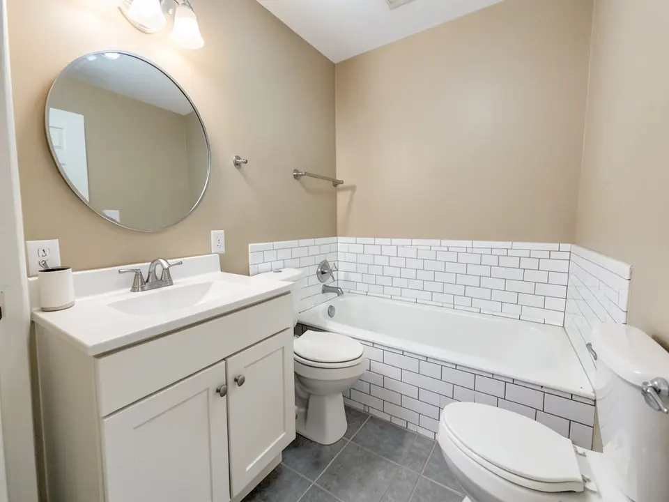Small bathroom with beige painted walls, a white vanity with a round mirror, a white bathtub with white subway tile and a gray tile floor, before
