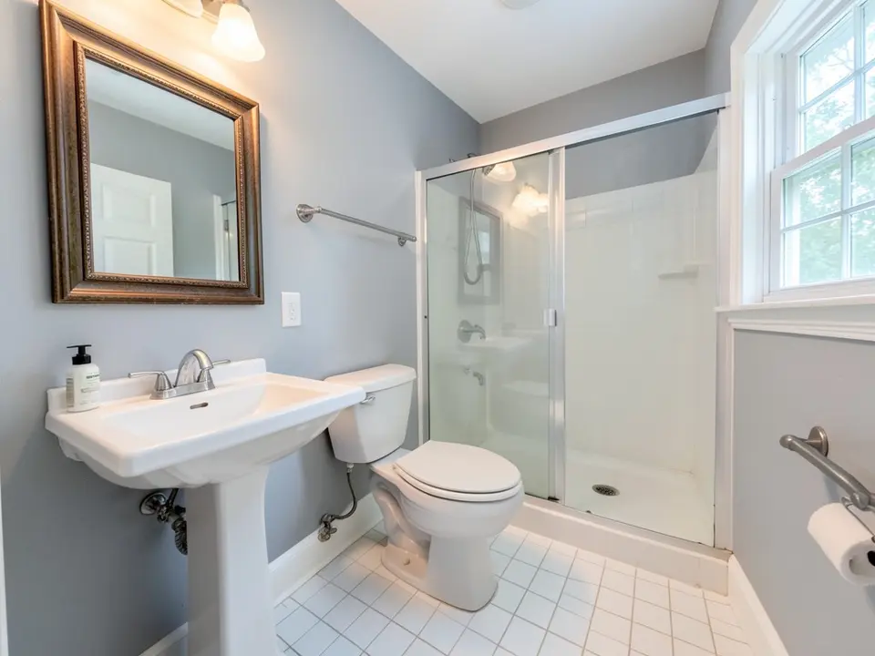 Bathroom with light gray painted walls, a white pedestal sink with a framed mirror, a shower with a glass door, a white tile floor and a window, before
