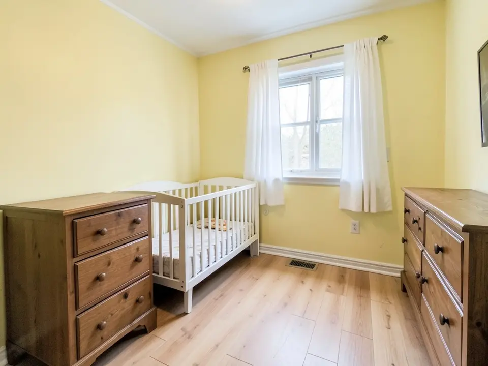 Small bedroom with pale yellow walls, a white crib, a wooden dresser, a window with white curtains and a light wood floor, before