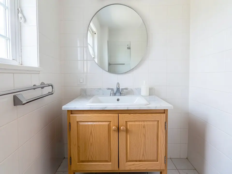 Bathroom with a light oak double-door vanity, white marble top, round mirror and white tile, before
