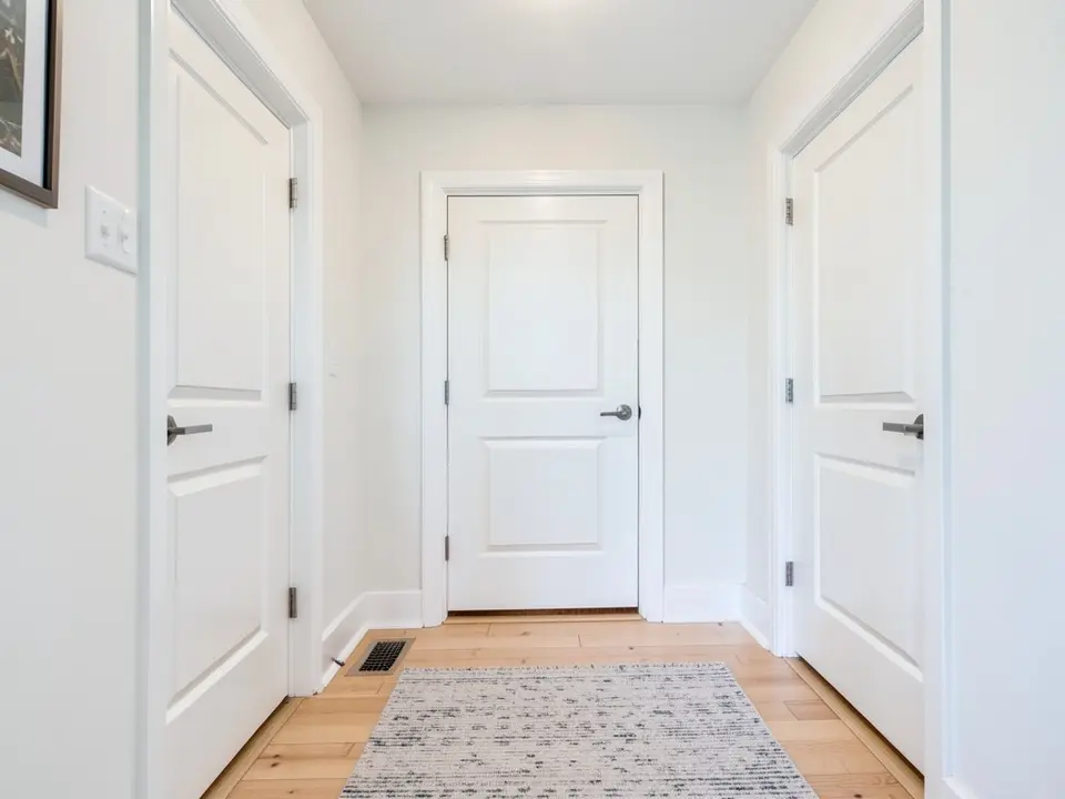 Hallway with white walls, white baseboards and casings, three closed white paneled doors with gray lever handles, a framed picture, a light wood floor and a patterned runner rug, before