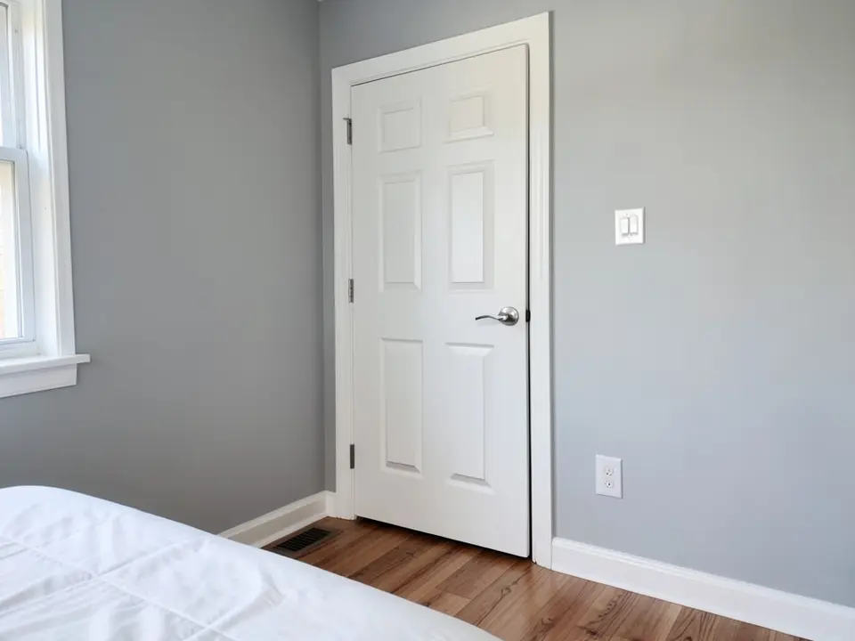Bedroom corner with light gray walls, white baseboards, a white casing around a six-panel white interior door with a silver lever handle, white bedding and a wood floor, before
