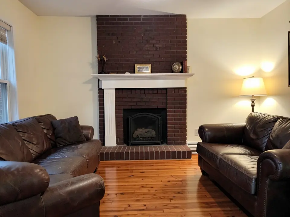 Living room with a dark red brick fireplace and hearth, a white mantel and white side pillars, framed art and decor on the mantel, cream walls, a table lamp, two brown leather sofas and a wood floor, before