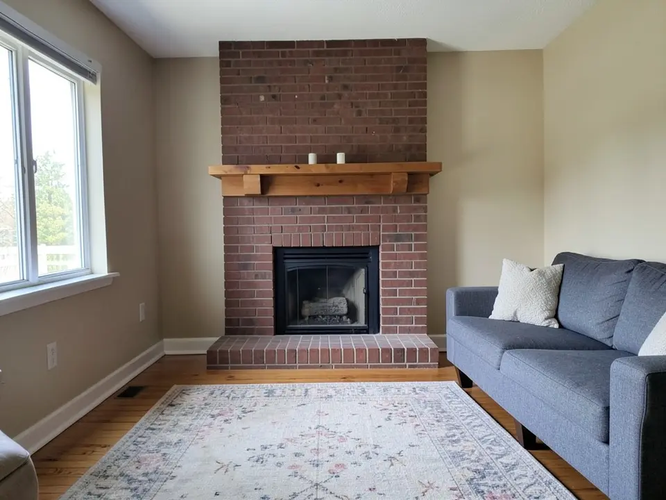 Living room corner with a red-brown brick fireplace and hearth, a rustic wooden mantel with two candles, beige walls, a window with blinds, a gray-blue sofa, a floral rug and a wood floor, before