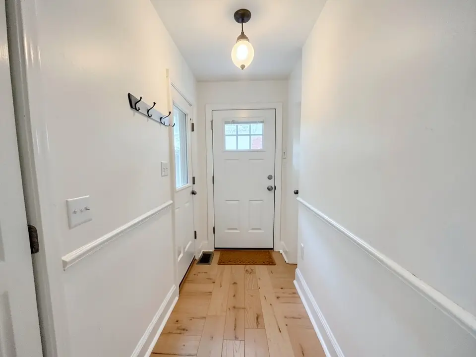 Entry hall with white walls, a white chair rail, a front door with a window, coat hooks, a pendant light, a doormat and a wood floor, before