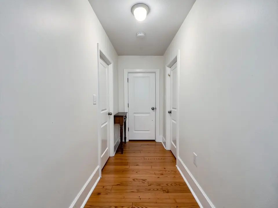 Narrow hallway with white walls, a wood floor, white doors, a small console table at the far end and a ceiling light, before