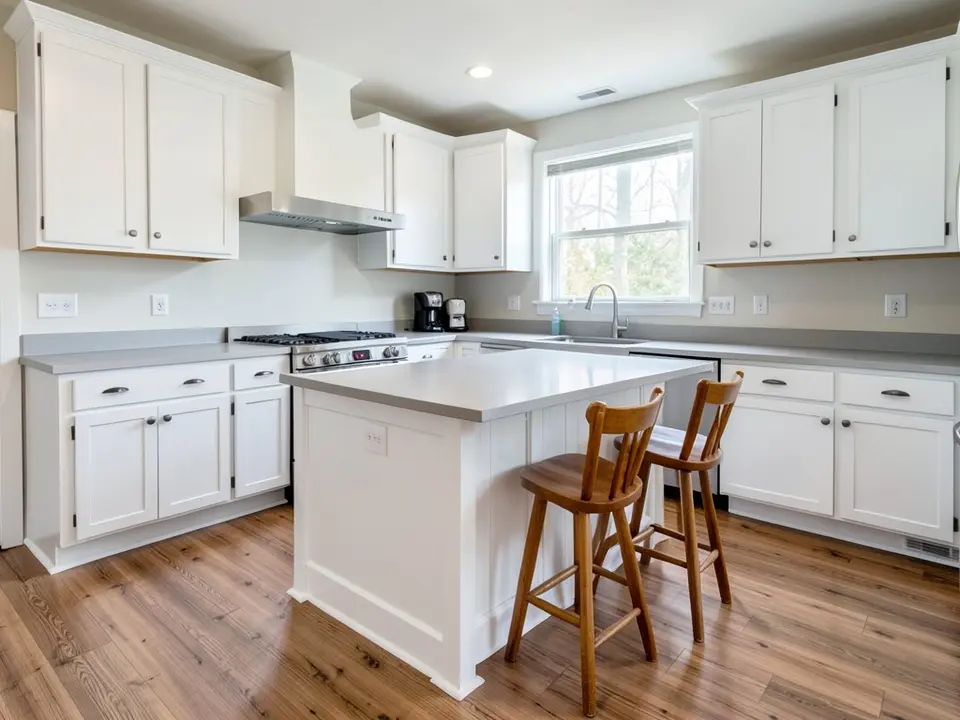 White shaker kitchen with white perimeter cabinets, a white kitchen island with two wooden bar stools, a light gray countertop and a wood floor, before