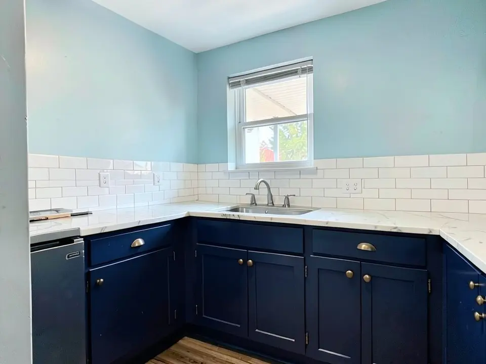 The same kitchen with the walls painted a pale soft blue, with the navy cabinets, brass pulls, countertop, backsplash and window kept
