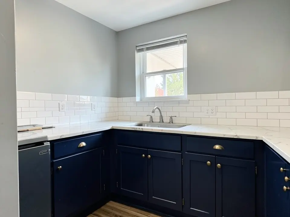 Kitchen with light gray walls, navy lower cabinets with brass pulls, a white marble countertop, a white tile backsplash and a window above the sink, before