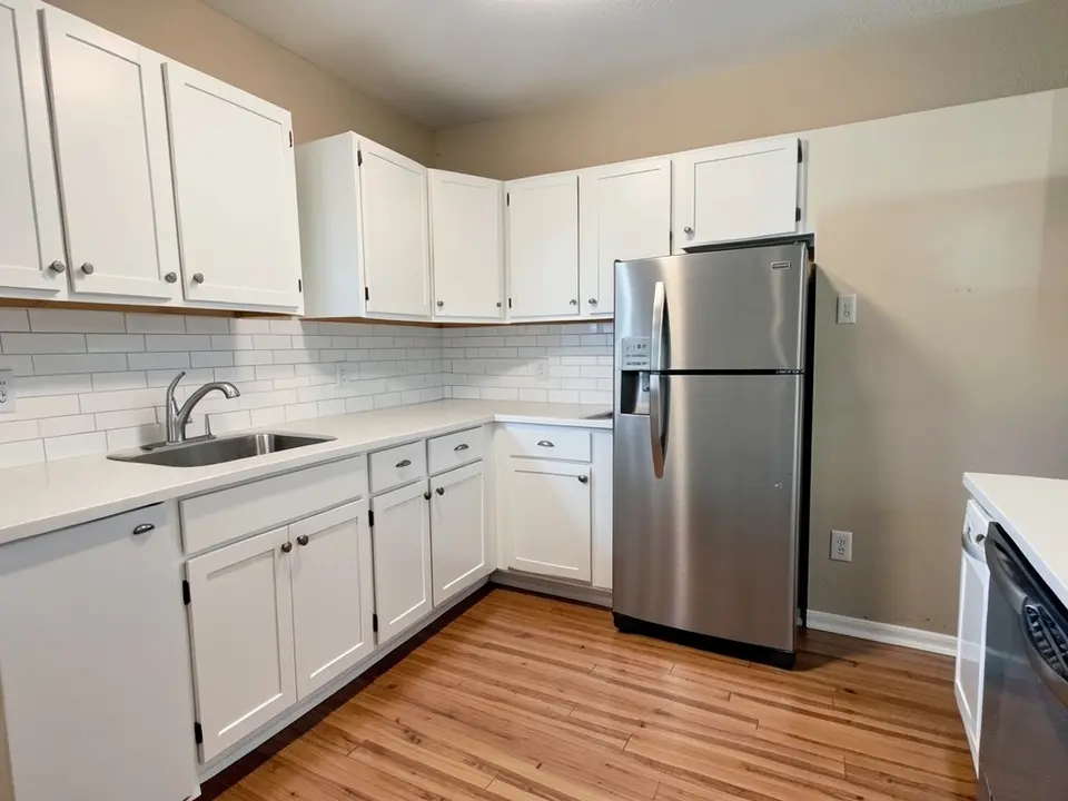Kitchen with beige walls, white shaker cabinets, a white countertop, a white subway tile backsplash, a stainless fridge and a wood floor, before