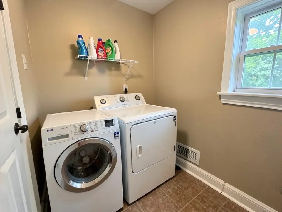 Laundry room with beige walls, a white washer and dryer side by side, a wire shelf with detergent bottles, a window with white trim and a brown floor, before