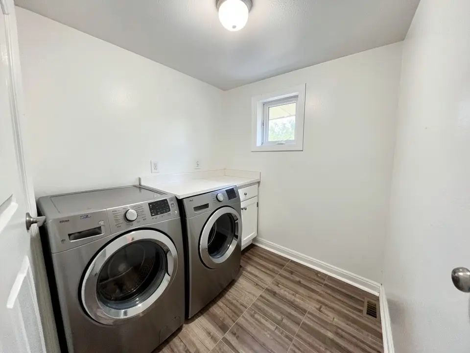 Laundry room with white walls, a gray metallic washer and dryer, a white countertop over a base cabinet, a small window and a wood-look floor, before