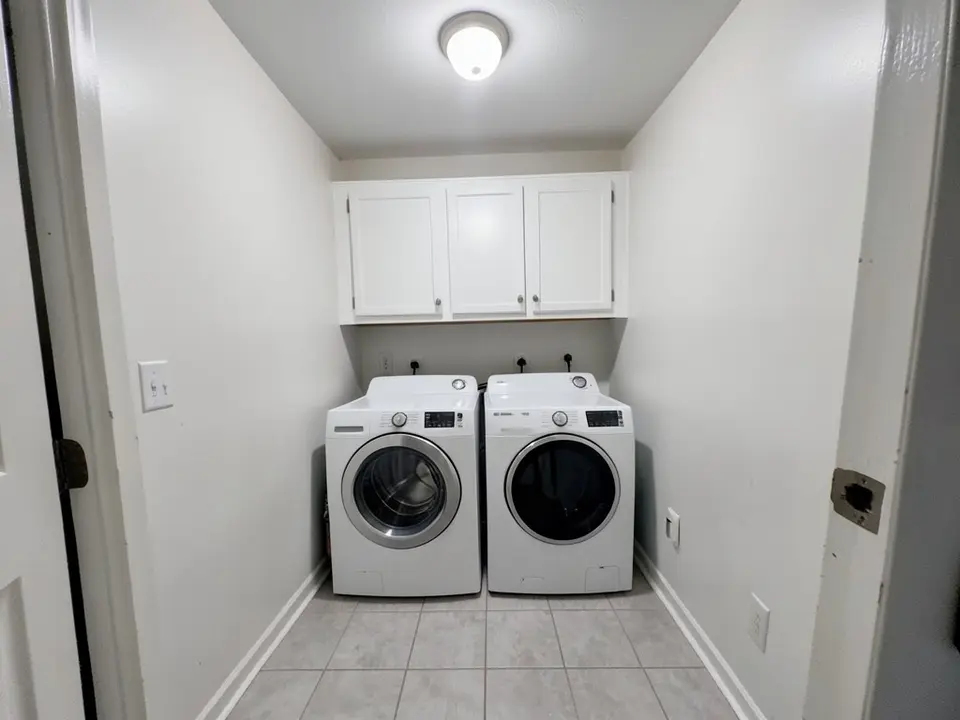Small laundry room with white walls, white front-load washer and dryer, white upper cabinets above them, light gray floor tile and a ceiling light, before