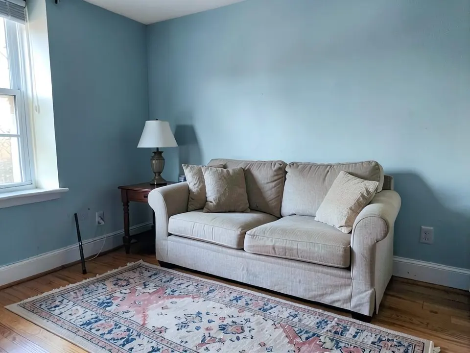 The same living room with the walls painted a muted soft blue, with the sofa, side table, lamp, rug and floor kept