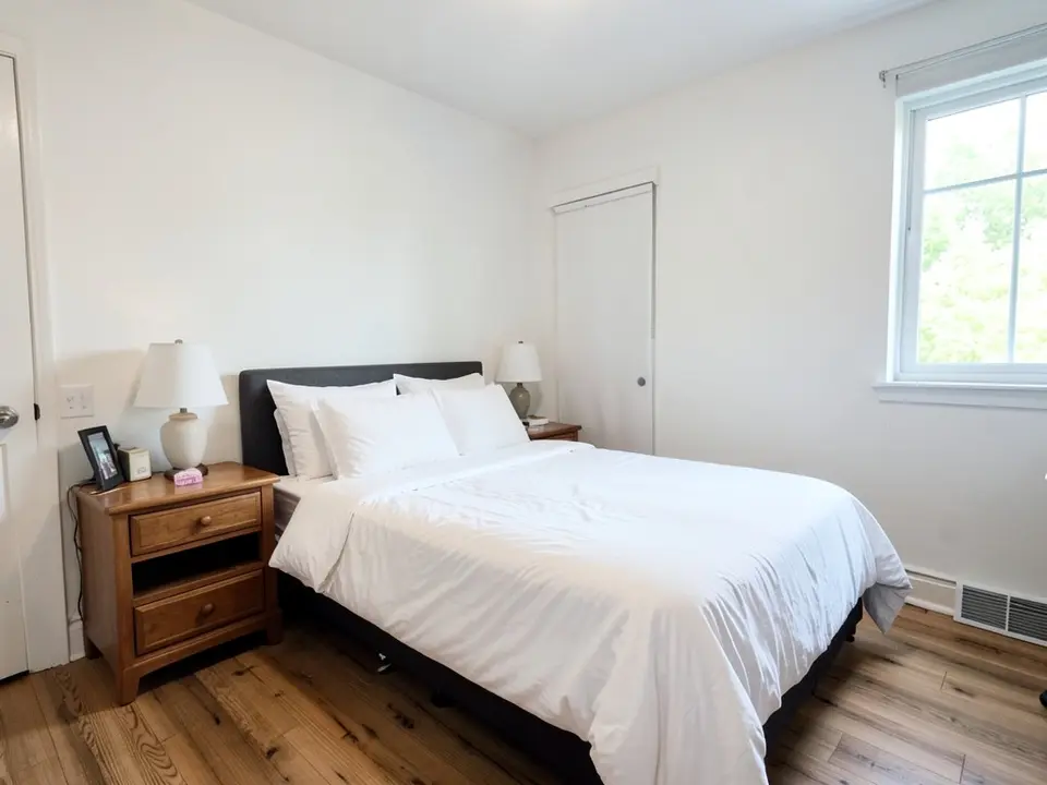 Bedroom with white walls, a bed with white bedding, a wooden nightstand with a lamp and a window, before
