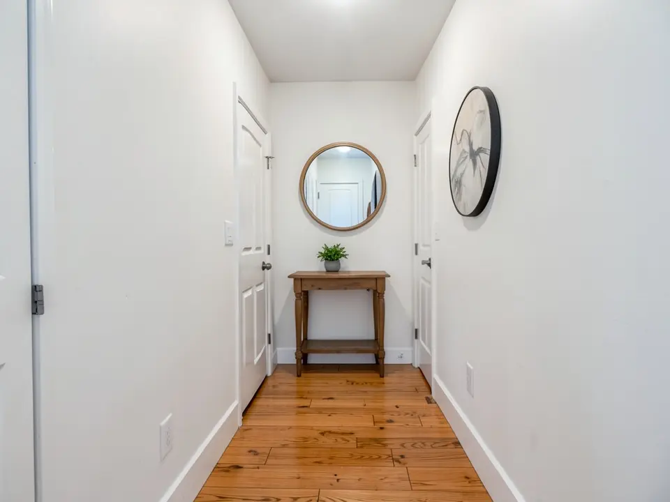Entryway hallway with white walls, a wooden console table with a plant, a round mirror and a wood floor, before