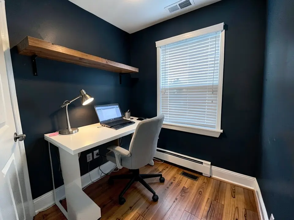 The same home office with the walls painted very dark navy-charcoal, with the desk, chair, shelf and blinds kept