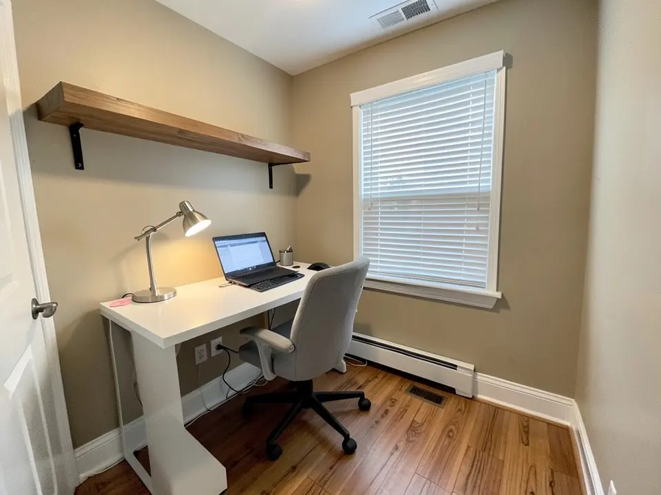 Home office with beige walls, a white desk with a laptop and a lamp, a gray chair, a floating wood shelf, a window with white blinds and a wood floor, before