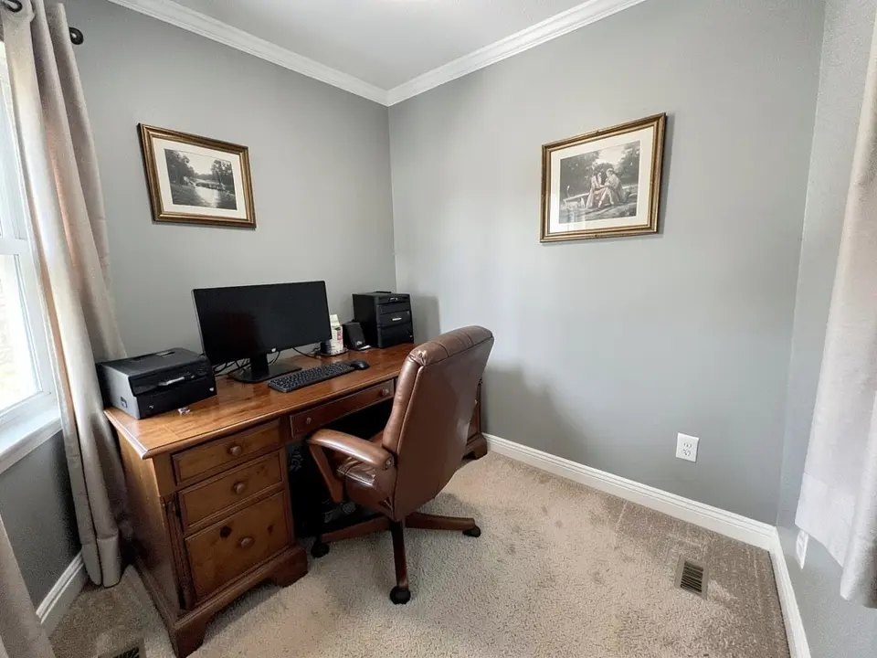 Home office with gray walls, white crown molding and baseboards, a wooden desk with a computer, a brown leather chair, framed pictures, curtains and beige carpet, before