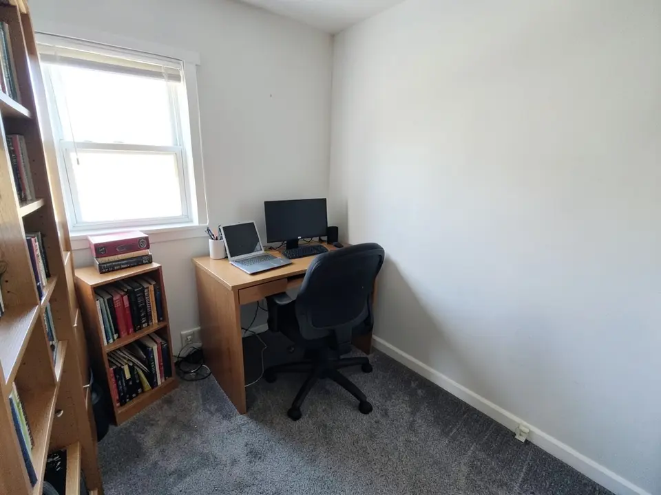 Small home office with white walls, a wooden desk with a laptop and monitor, a black office chair, bookshelves, a window and gray carpet, before