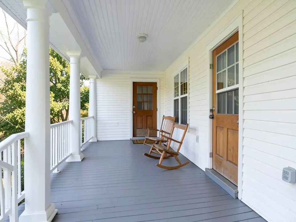 Front porch of a white clapboard house with white columns, a gray wood floor, a white beadboard ceiling, a wooden front door and a rocking chair, before