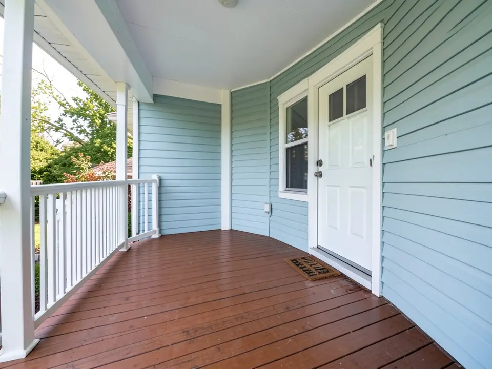 Covered porch with white railings and posts, a stained reddish-brown wood floor, light blue siding and a white front door, before