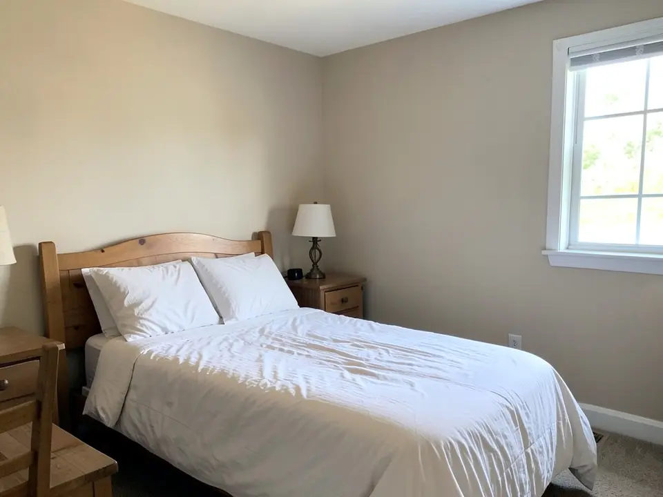 Bedroom with light beige walls, a bed with white bedding, a wooden nightstand with a lamp and a window, before