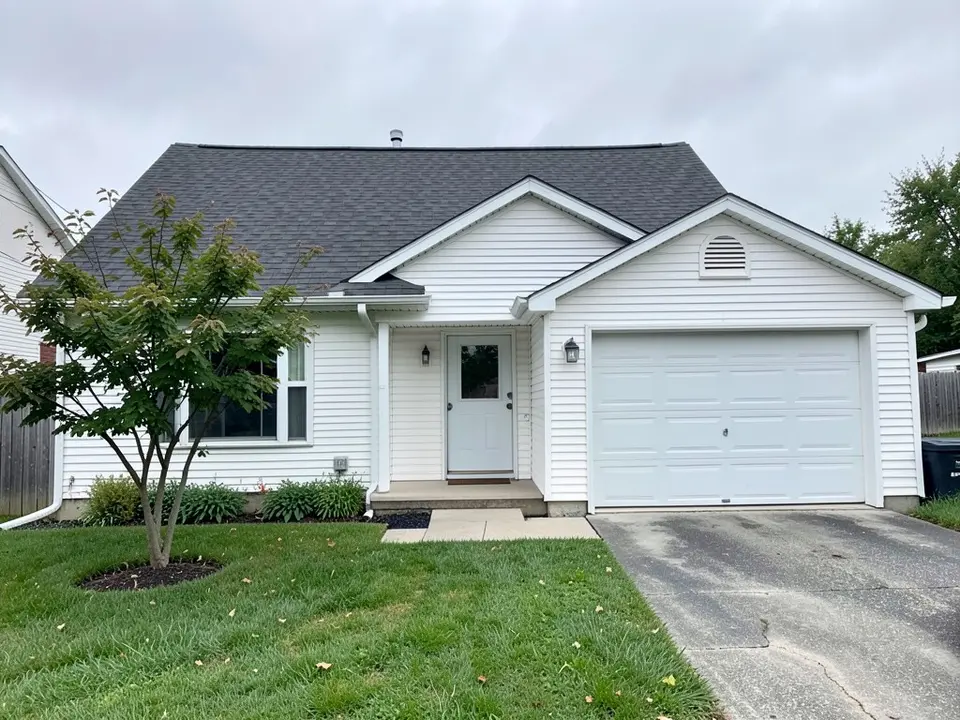 One-story house with white vinyl siding, a white garage door, a dark gray roof and a small tree, before