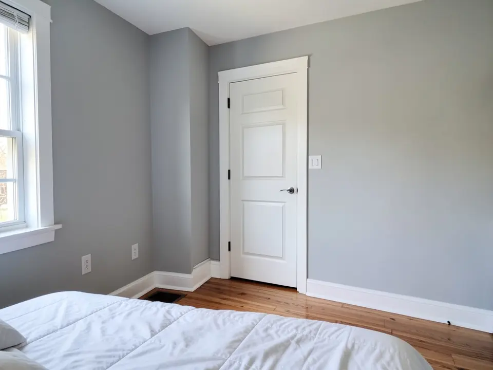 Bedroom with light gray walls, white baseboards, a white paneled door with white casing and a white window frame, before